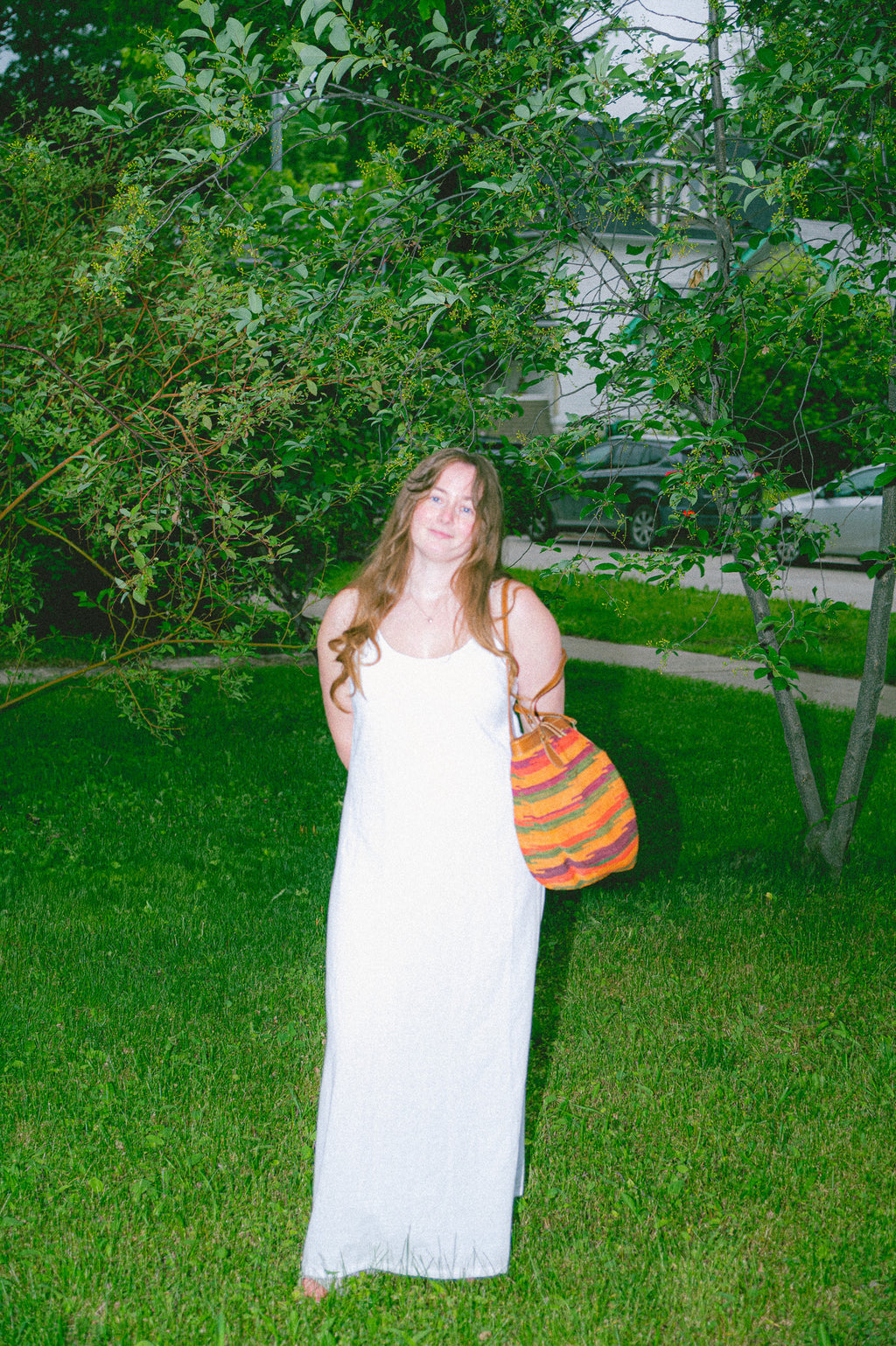 Woman in a true white dress holding a colorful bag in a grassy area with trees and houses in the background.