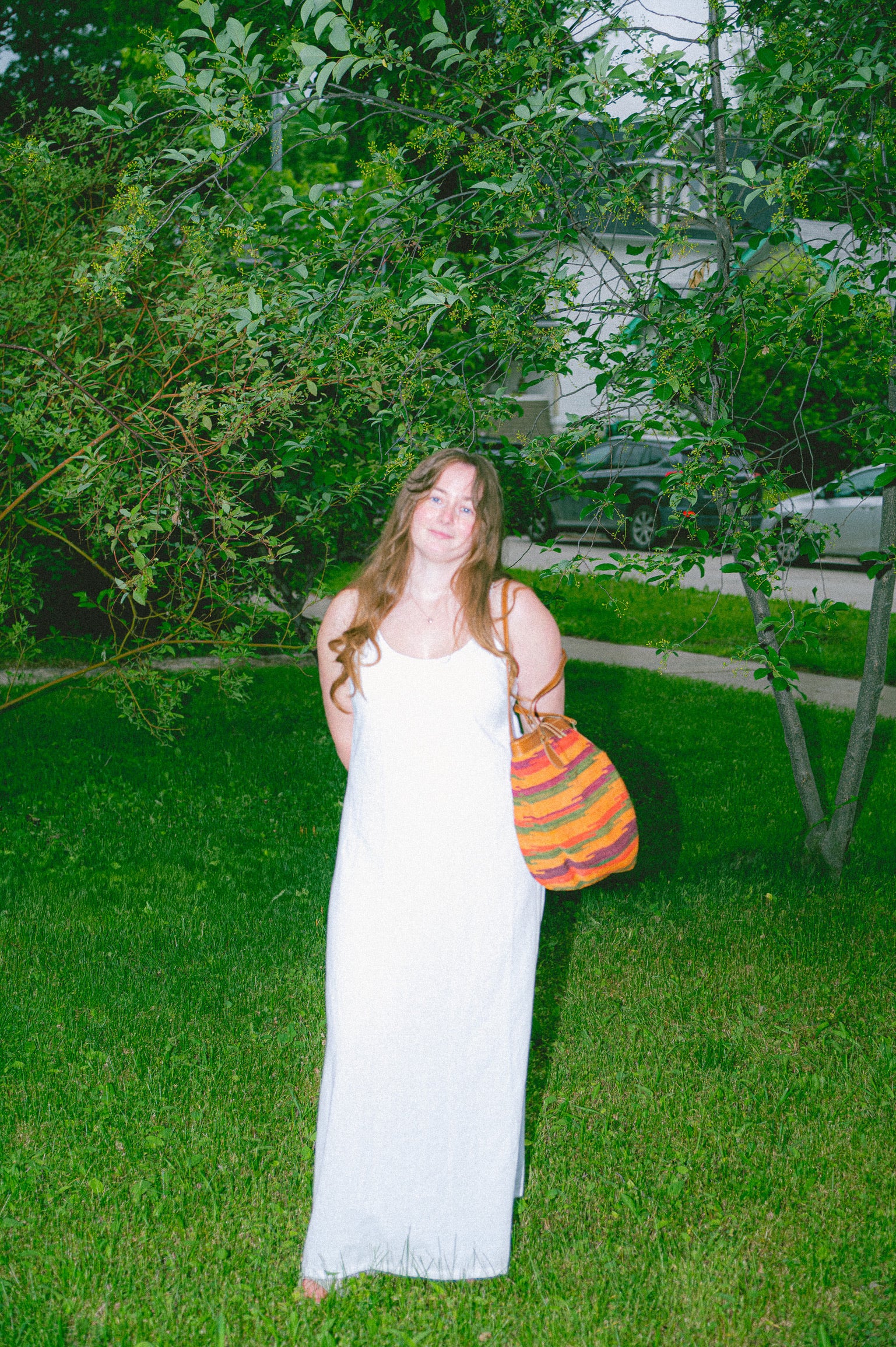Woman in a true white dress holding a colorful bag in a grassy area with trees and houses in the background.