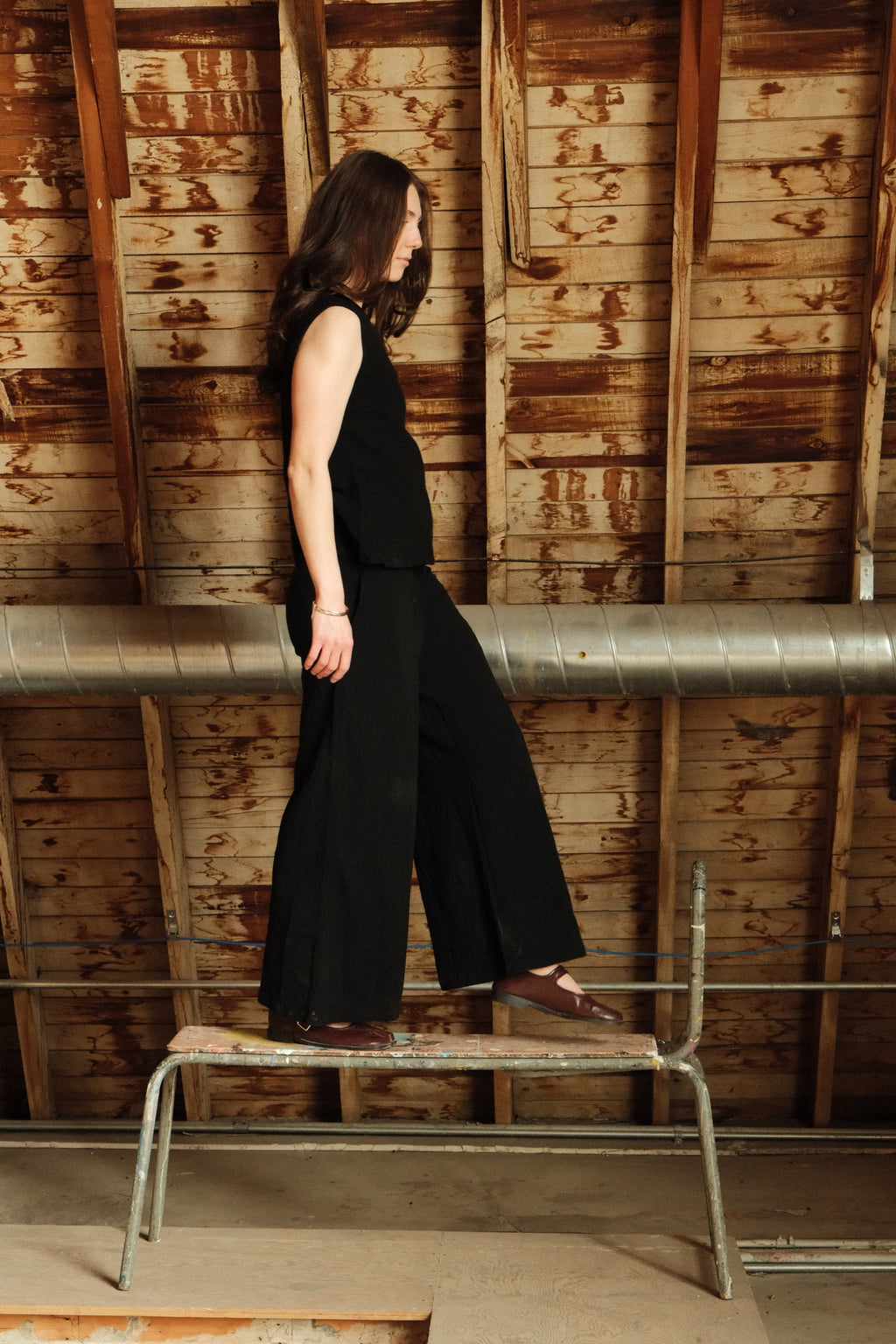 Woman in a black outfit standing on a metal stool against a wooden wall.