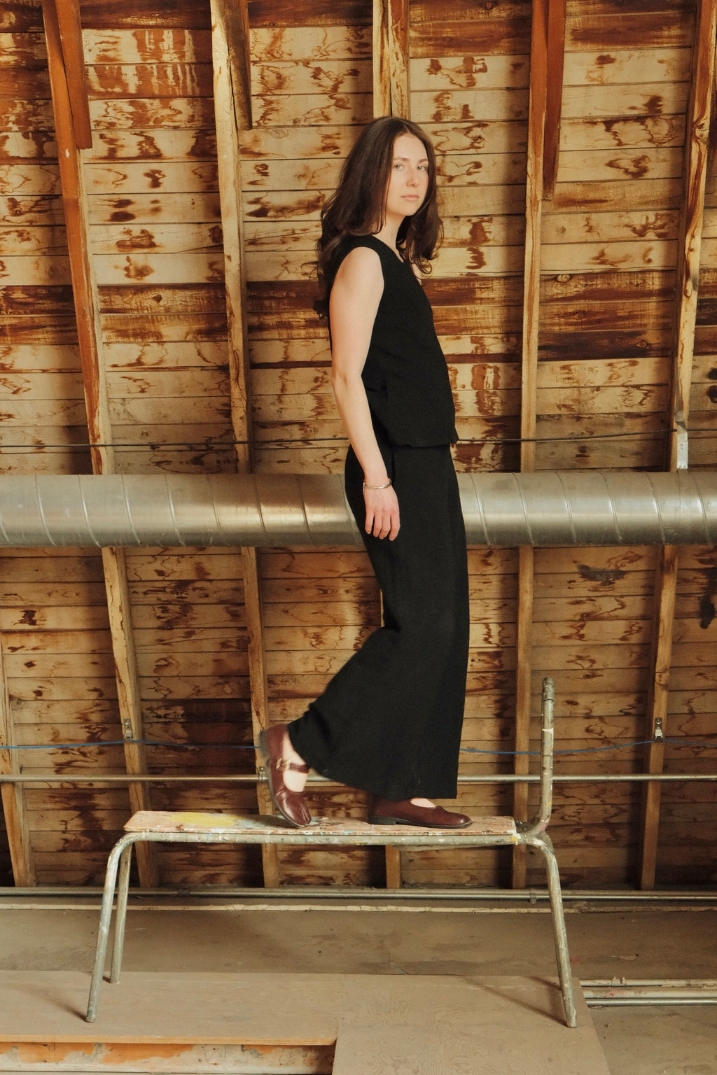 Woman in a black outfit standing on a metal bench against a wooden wall.