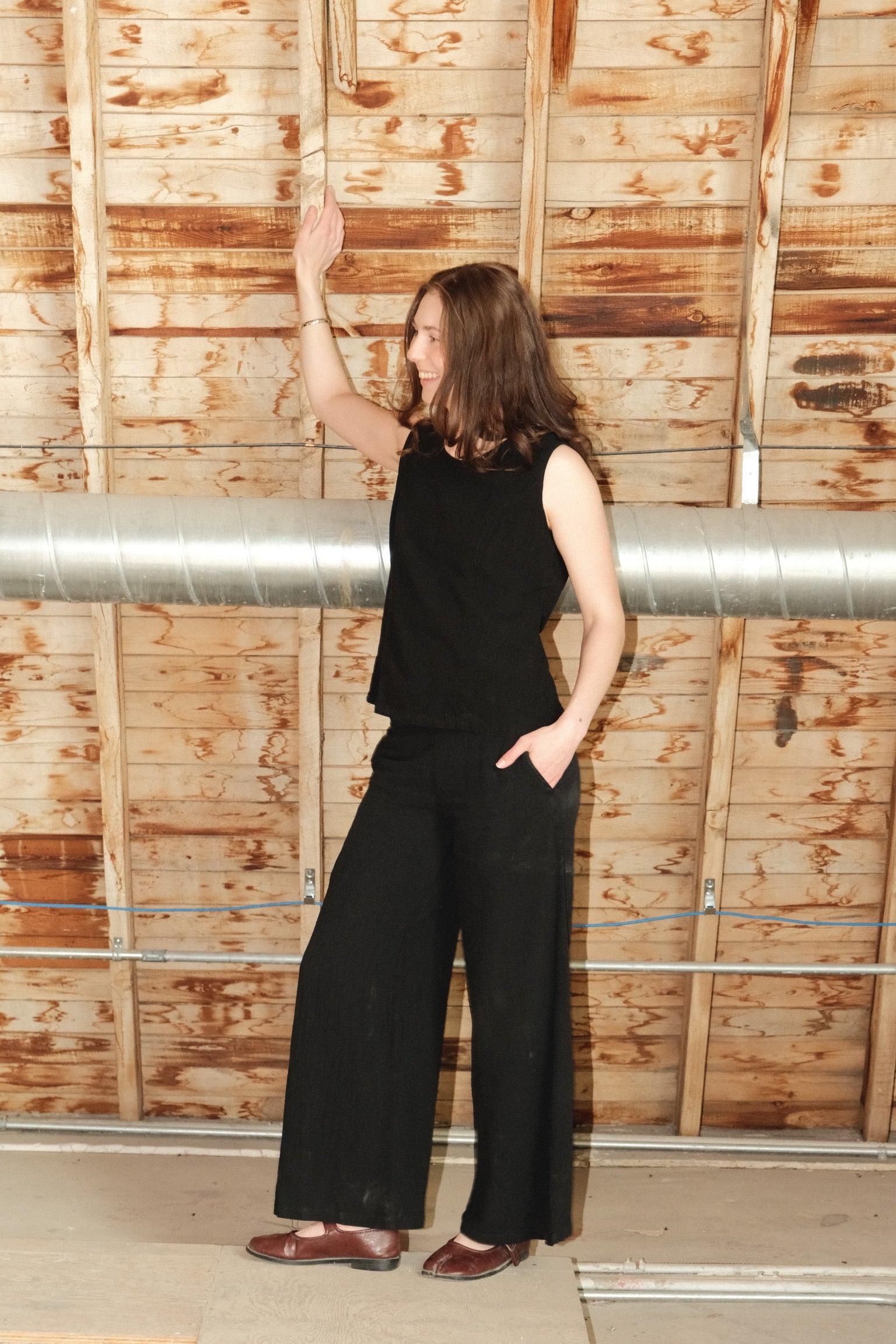 Woman in black sleeveless top and wide-leg pants standing against a wooden wall.